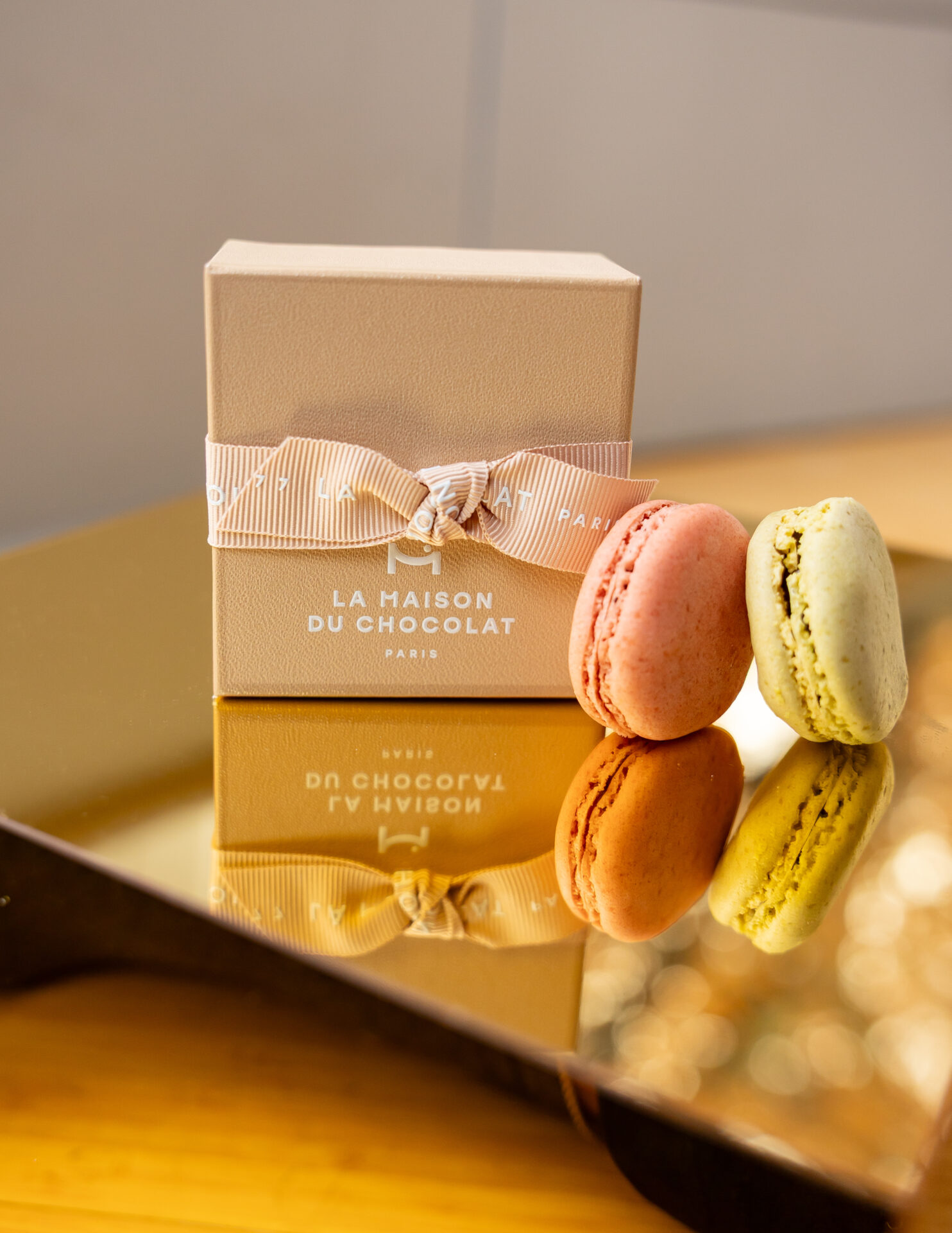 La Maison du Chocolat gift box tied with pink ribbon beside pink and pistachio macarons on a gold mirrored tray