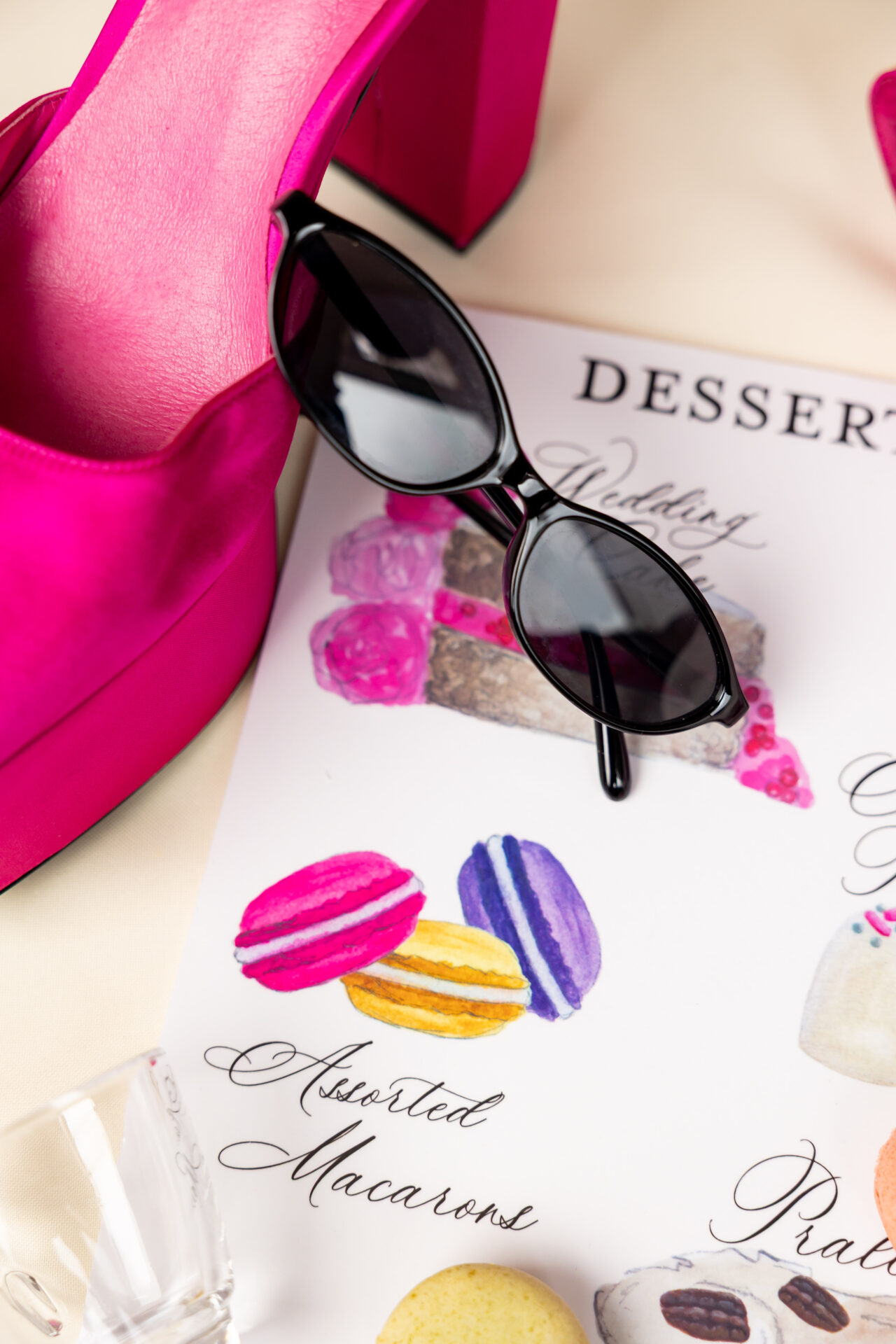 Close-up of black sunglasses resting on a hand-painted dessert menu beside a hot pink platform shoe