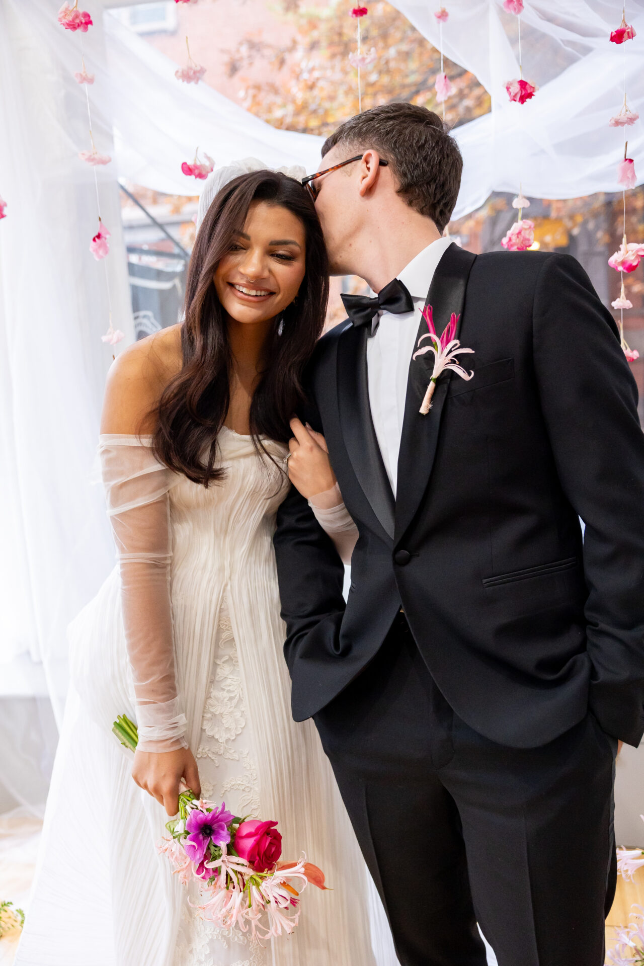 Groom in a black tuxedo kissing the smiling bride's forehead under hanging pink flowers at Atrium82 in Brooklyn