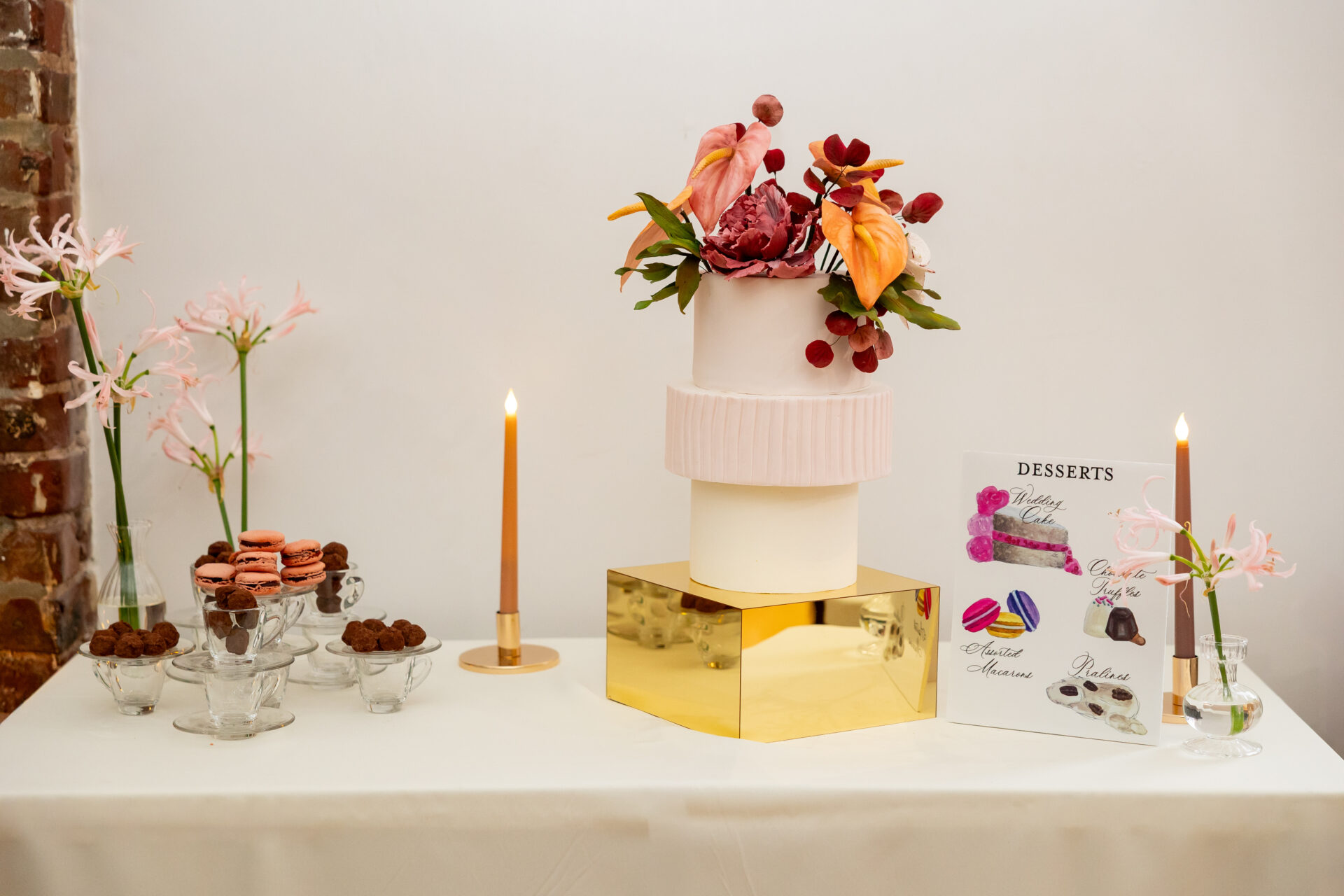 Dessert table with a three-tier blush wedding cake on a gold mirrored block, macarons, truffles, taper candles, and a watercolor dessert sign