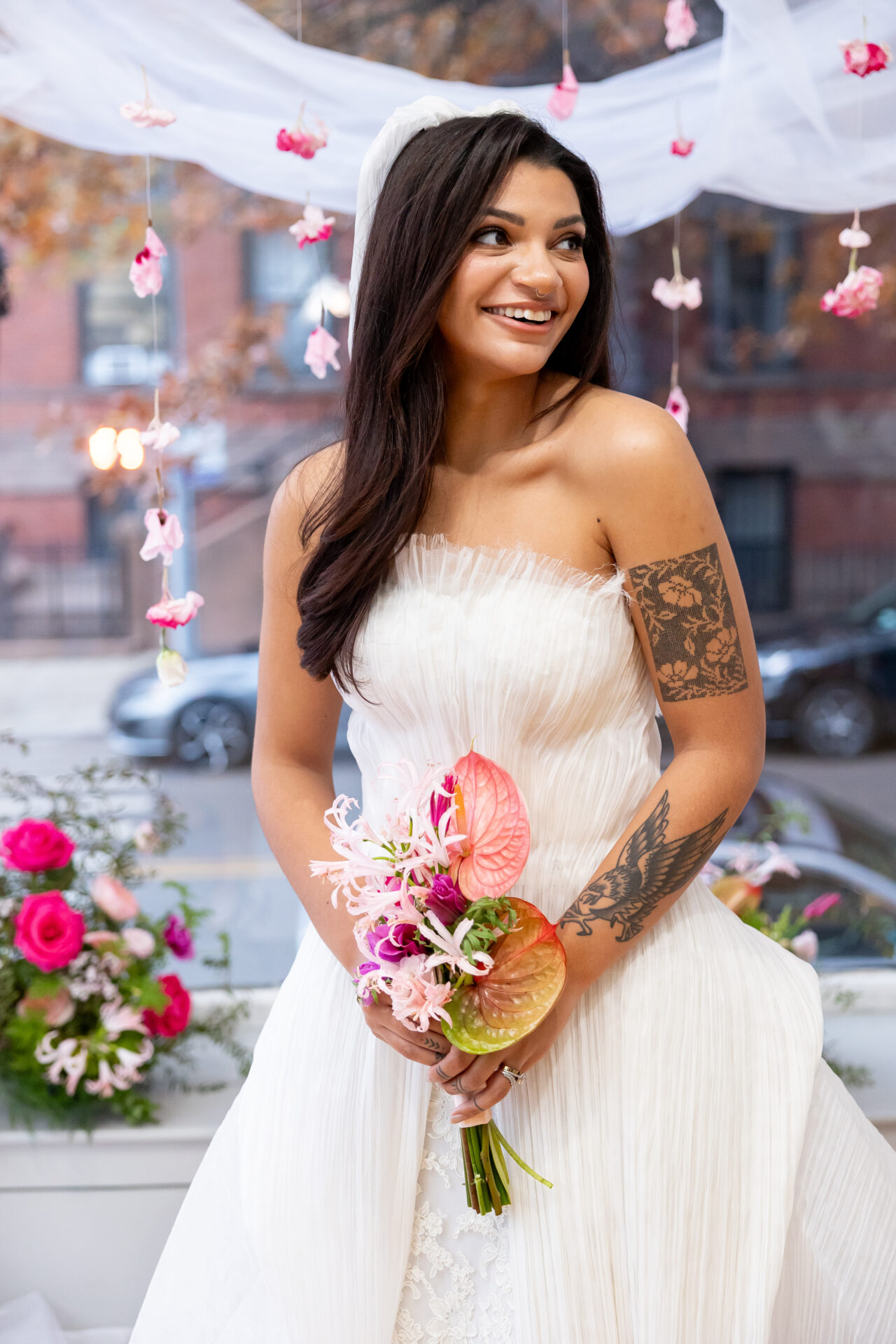 Smiling bride in a strapless pleated gown holding an anthurium and nerine bouquet under hanging pink blooms