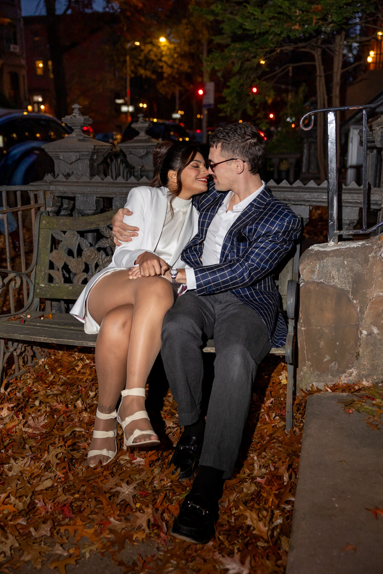 Couple on an iron bench outside a Brooklyn brownstone at night, surrounded by fallen autumn leaves